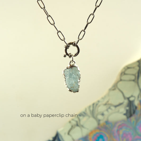 A silver paperclip chain necklace with a raw, irregular-cut light blue aquamarine gemstone pendant