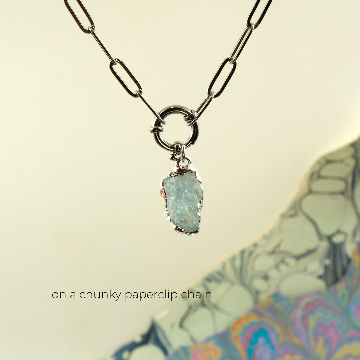 A silver paperclip chain necklace with a raw, unpolished light blue-green aquamarine gemstone pendant