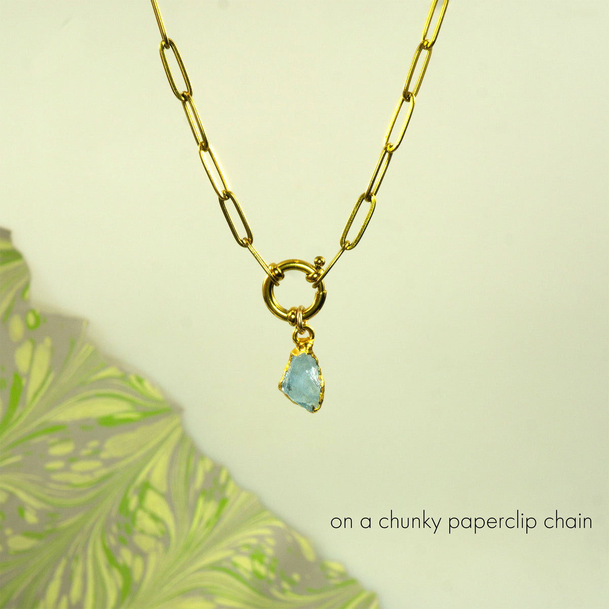 A gold paperclip chain necklace with a gold ring clasp and a small aquamarine teardrop gemstone pendant