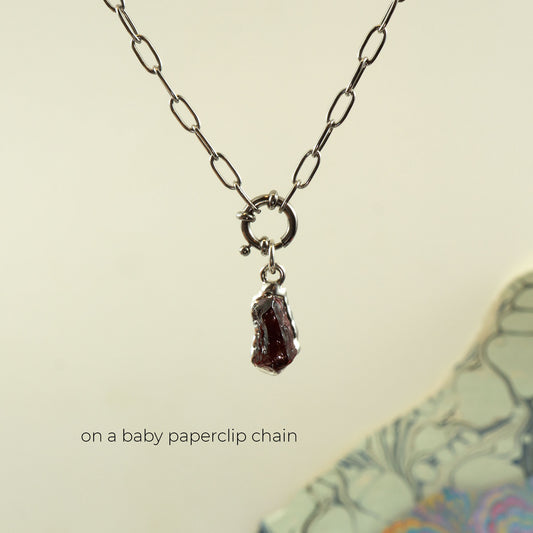 A silver paperclip chain necklace with a small ring clasp and a dangling rough dark red garnet crystal pendant