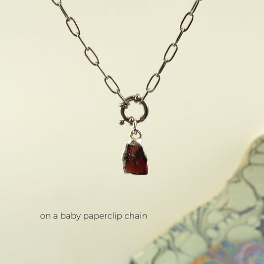 A silver paperclip chain necklace with a small ring clasp and a raw, irregular dark red garnet-pendant gemstone pendant