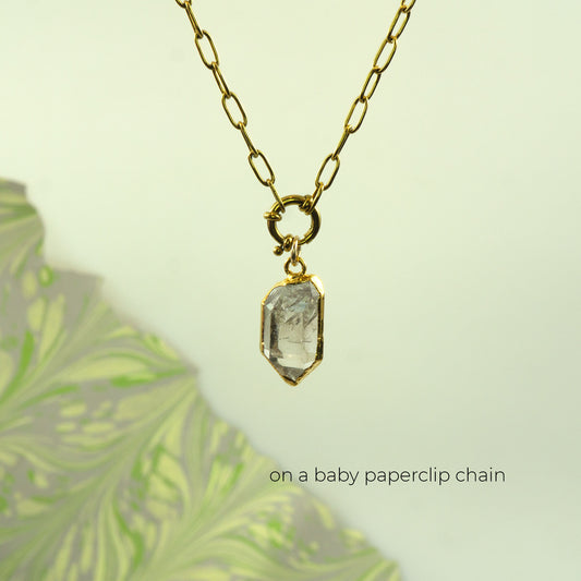 A gold paperclip chain necklace with a small gold clasp holding a clear faceted Herkimer Diamond crystal pendant
