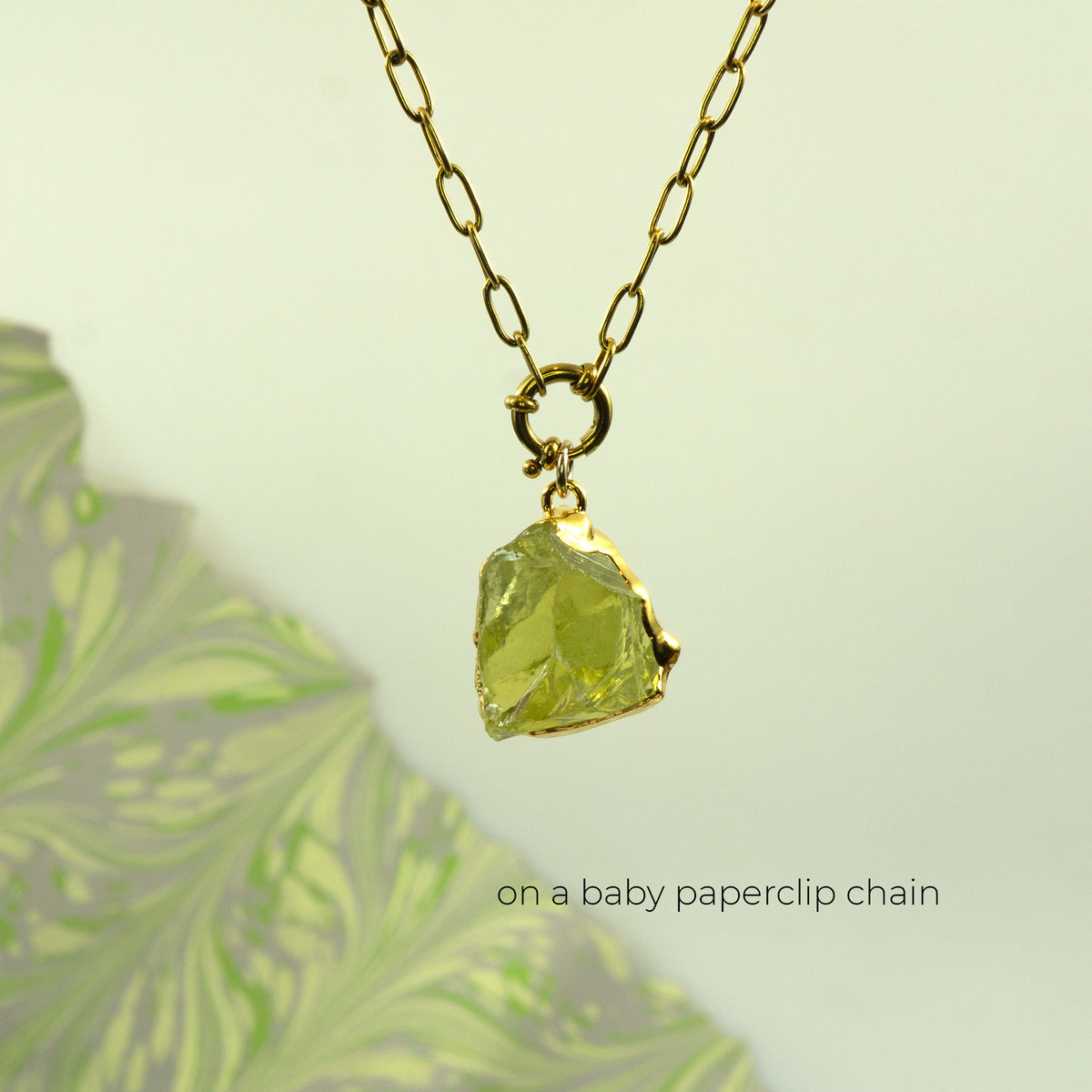 A gold necklace with a raw lemon quartz, faceted yellow-green gemstone pendant, suspended from a gold paperclip chain.