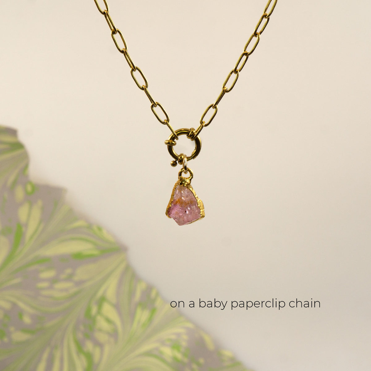 A gold paperclip chain necklace with a small gold ring clasp holding a rough Pink Tourmaline gemstone pendant,