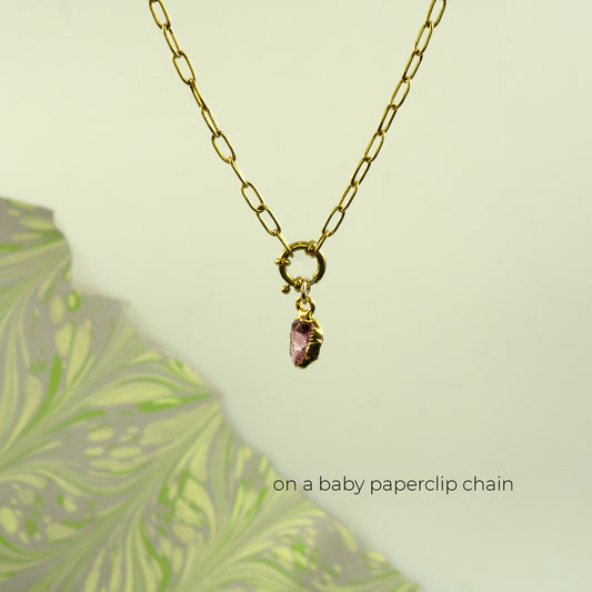 A gold paperclip chain necklace with a small gold clasp holding a dangling pink rhodlite garnet gemstone pendant