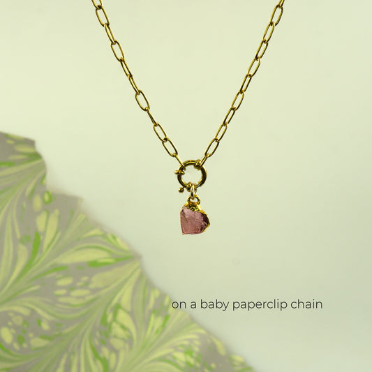 A gold paperclip chain necklace with a small ring clasp and a dangling raw pink rhodolite garnet stone charm