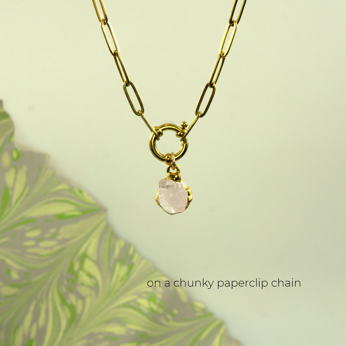A gold paperclip chain necklace with a small ring clasp and a raw, irregular pale pink raw rose quartz gemstone pendant
