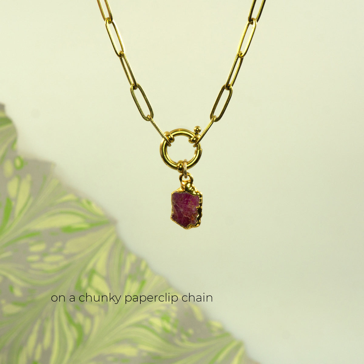 A gold paperclip chain necklace with a small ring connector and a raw, faceted ruby gemstone pendant