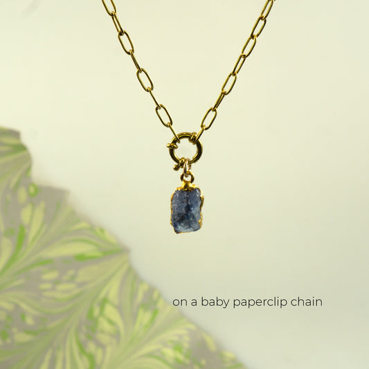 A gold chain necklace with a raw, oblong-shaped blue-gray tanzanite stone pendant