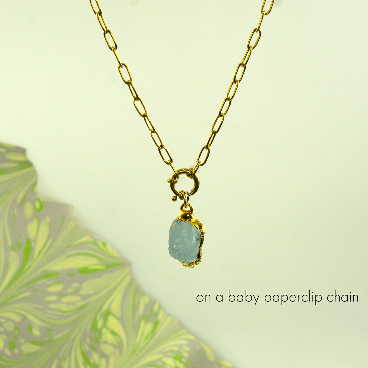 A gold paperclip chain necklace with a small gold clasp holding a rough, aquamarine gemstone pendant