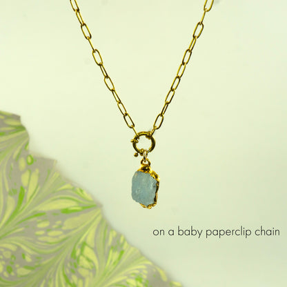 A gold paperclip chain necklace with a small gold clasp holding a rough, aquamarine gemstone pendant