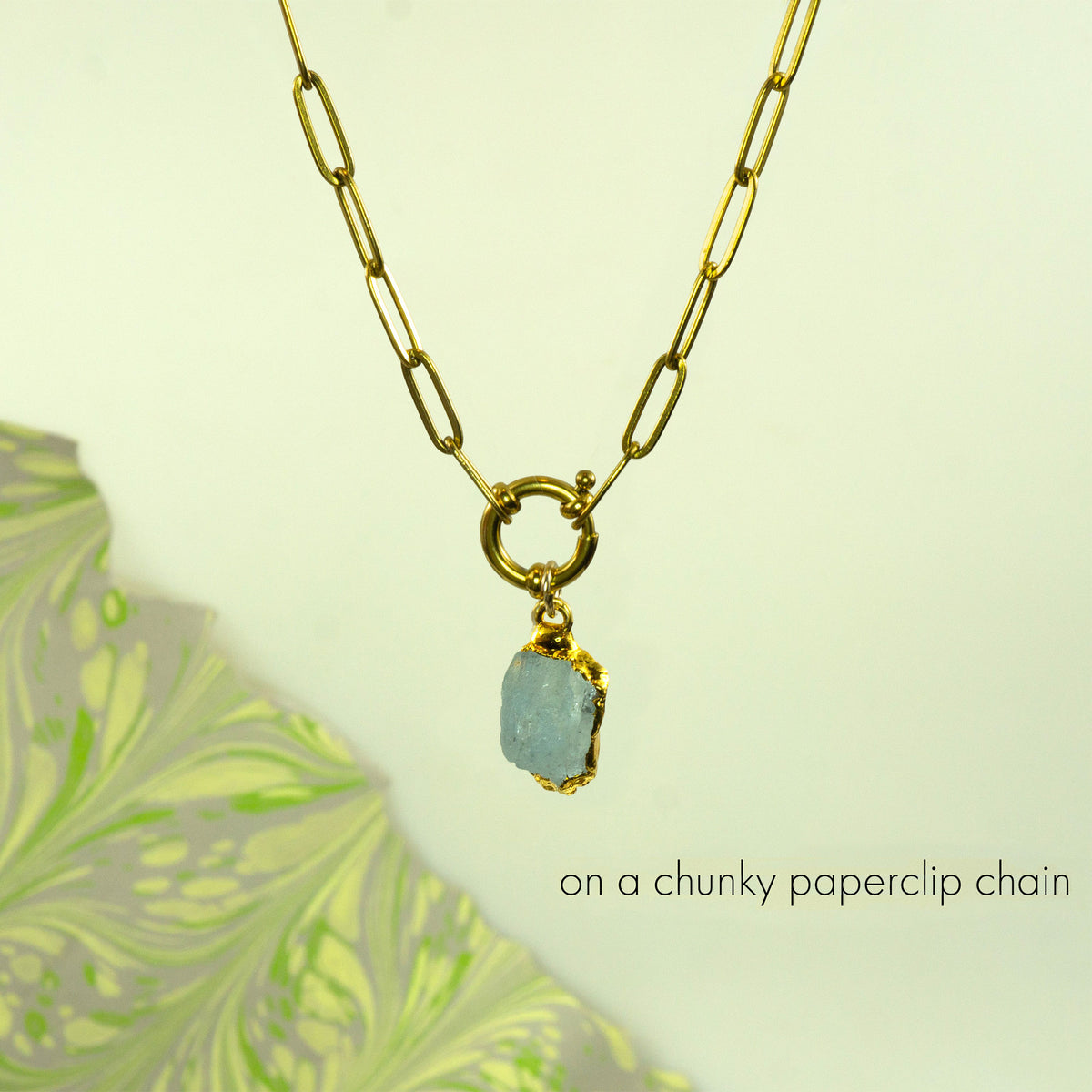 A gold paperclip chain necklace with a small ring clasp holding a raw, light blue aquamarine stone pendant.