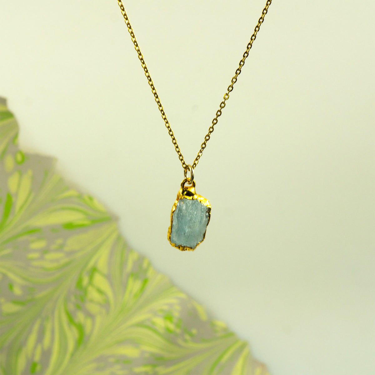 A gold chain necklace with a raw, irregular light aquamarine gemstone pendant