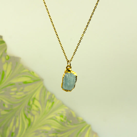 A gold chain necklace with a raw, irregular light aquamarine gemstone pendant