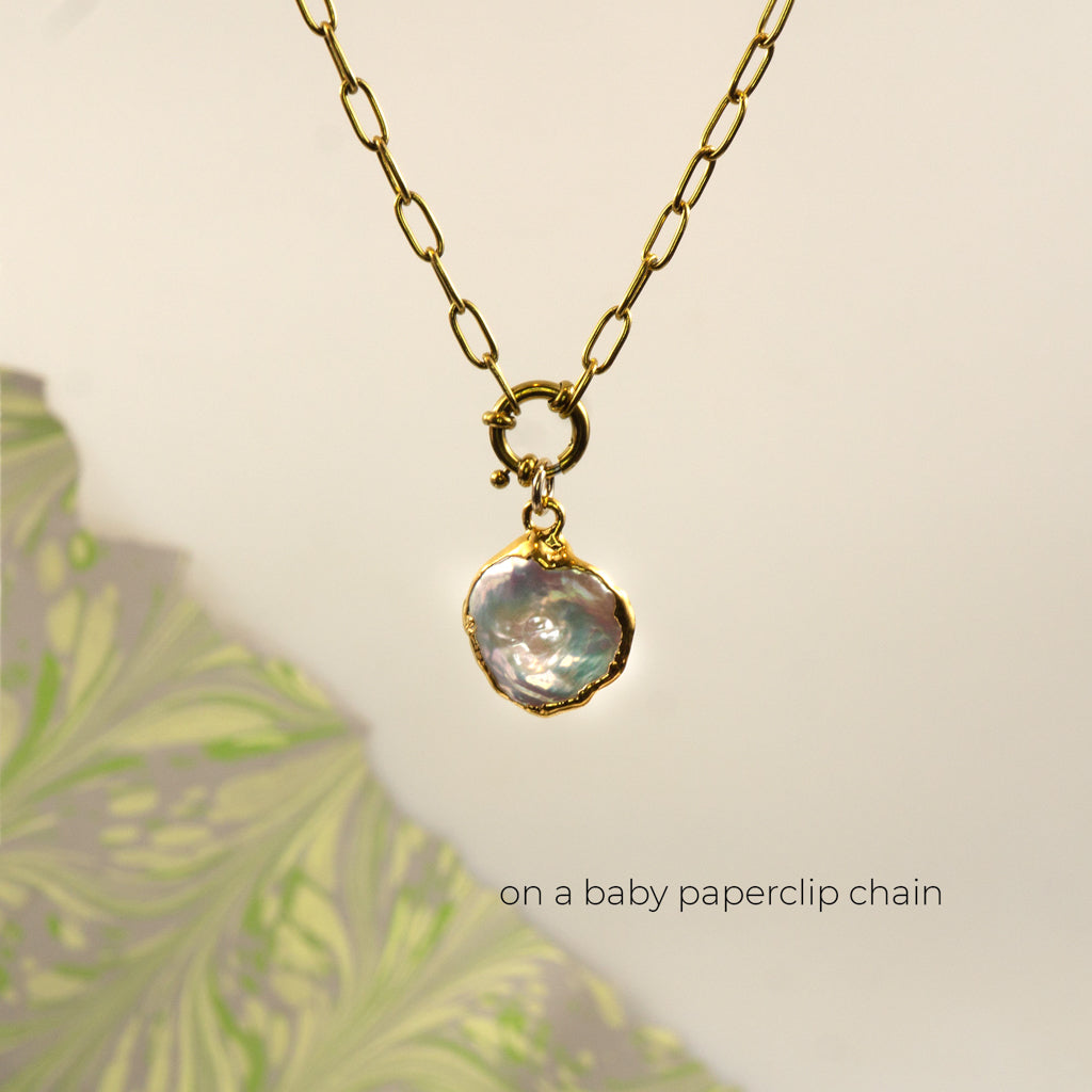A gold paperclip chain necklace with a gold clasp holding an irregular, iridescent pearl pendant