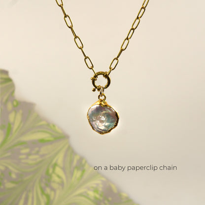 A gold paperclip chain necklace with a gold clasp holding an irregular, iridescent pearl pendant