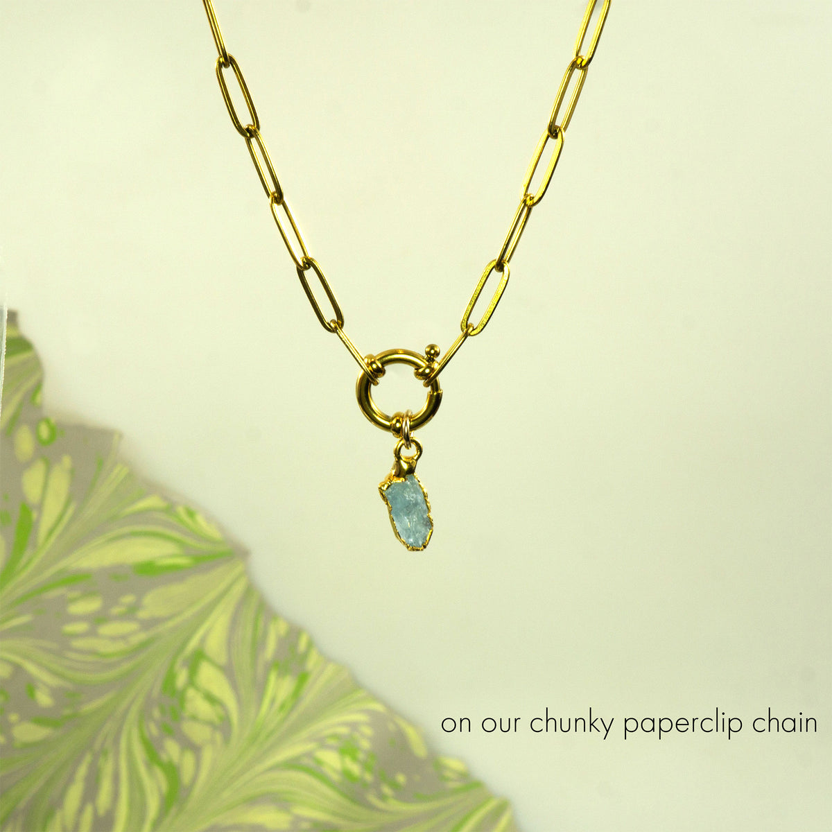 A gold paperclip chain necklace with a toggle clasp and a small light aquamarine gemstone pendant