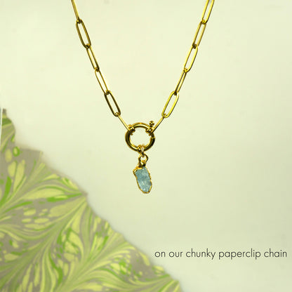 A gold paperclip chain necklace with a toggle clasp and a small light aquamarine gemstone pendant