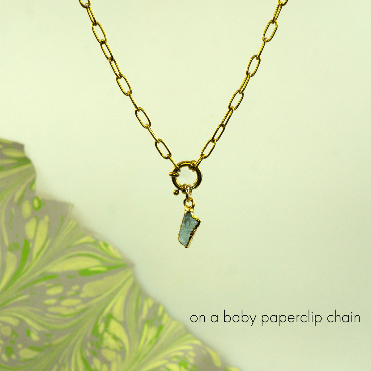 A gold paperclip chain necklace with a small gold clasp and a dangling raw, irregular aquamarine stone pendant