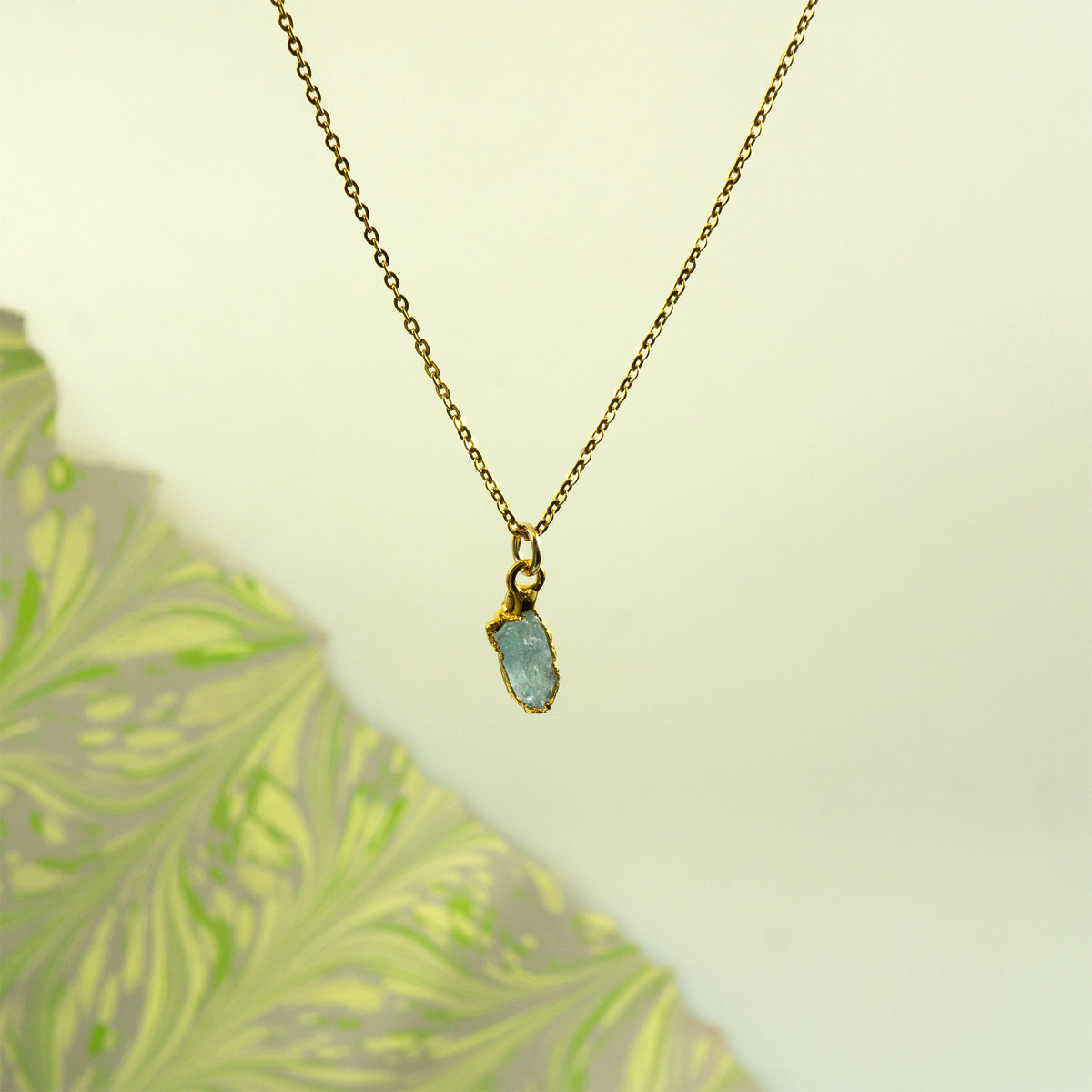A delicate gold chain necklace with a small gold bezel holding a flat, irregular light blue aquamarine gemstone pendant