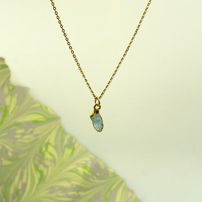 A delicate gold chain necklace with a small gold bezel holding a flat, irregular light blue aquamarine gemstone pendant