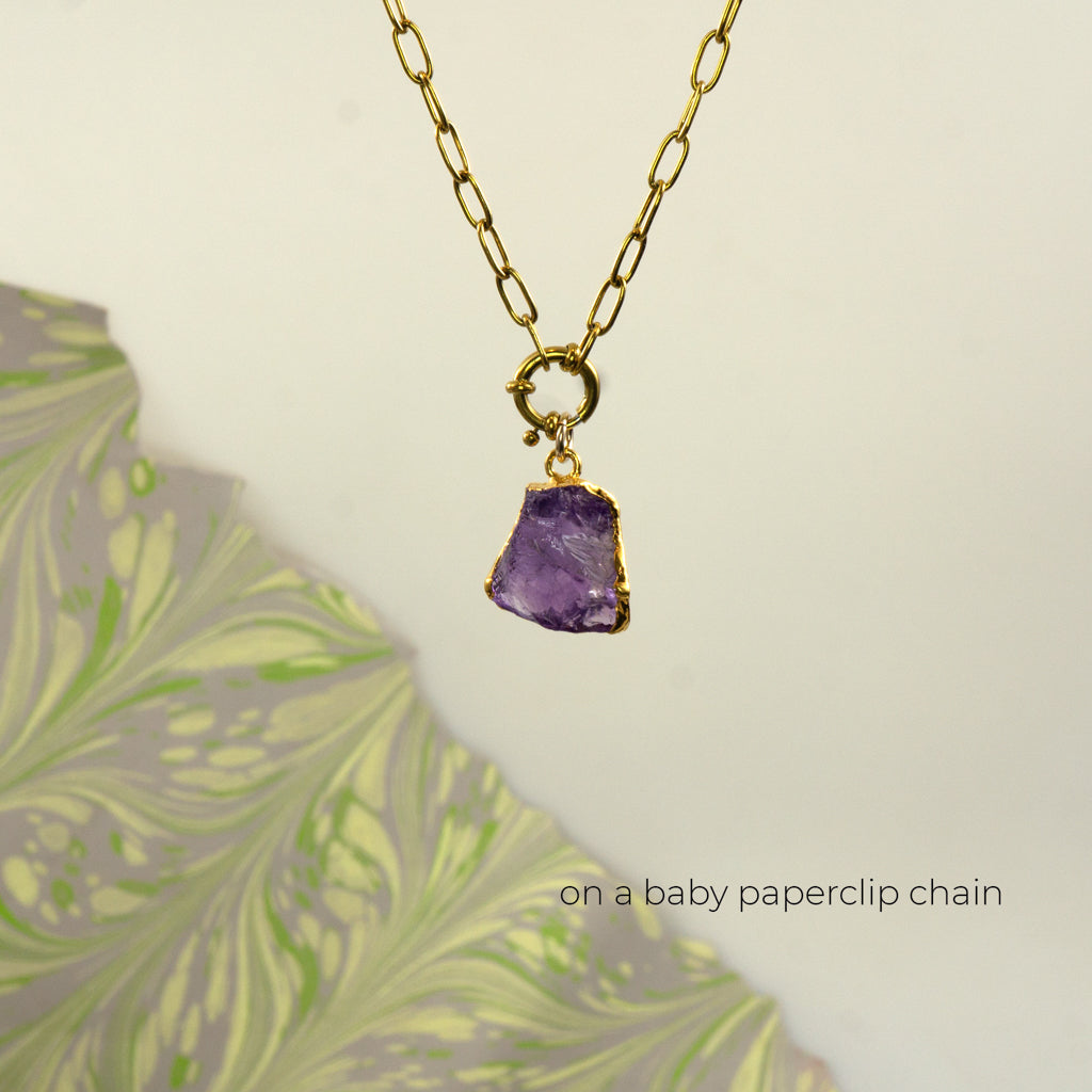 A gold paperclip chain necklace with a raw purple amethyst stone pendant set in a gold bezel 