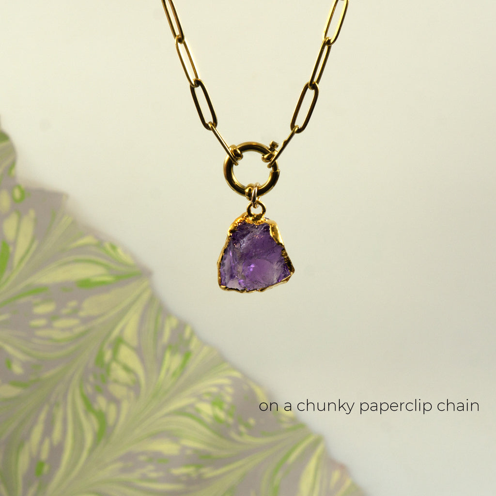 A gold paperclip chain necklace with a raw purple amethyst gemstone pendant set in gold