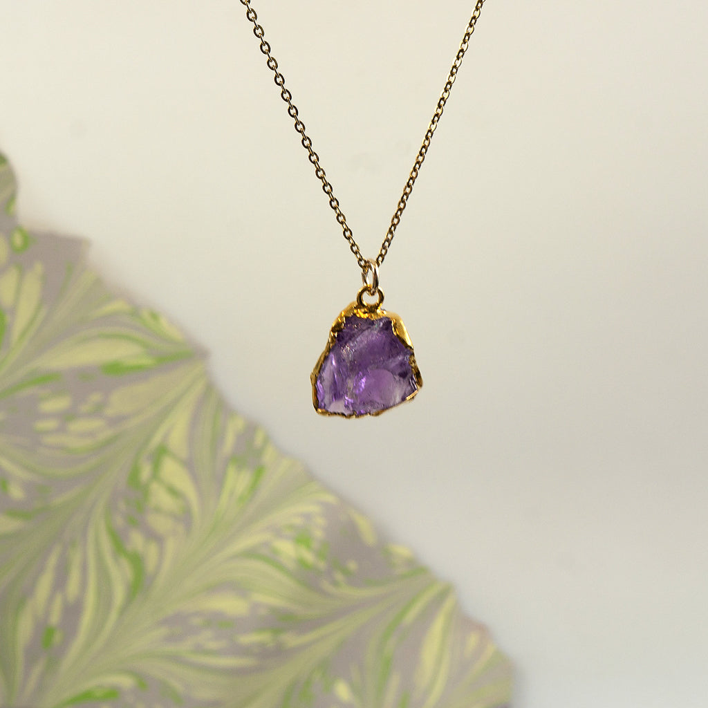 A raw amethyst crystal pendant necklace with a gold bezel setting on a delicate gold chain.