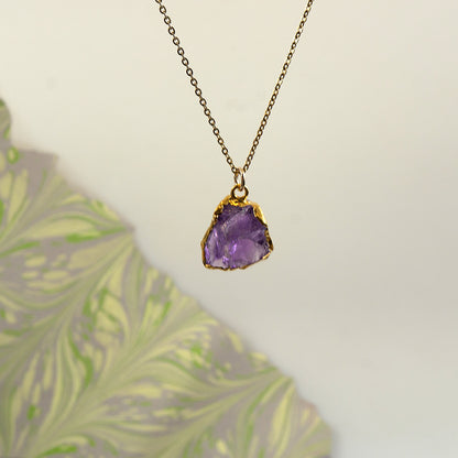 A raw amethyst crystal pendant necklace with a gold bezel setting on a delicate gold chain.