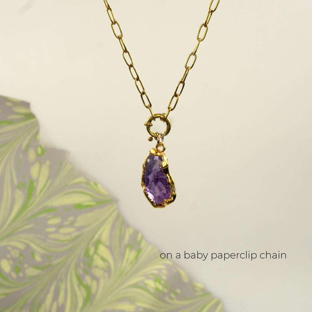 A gold paperclip chain necklace with a gold clasp holding a raw Amethyst pendant.