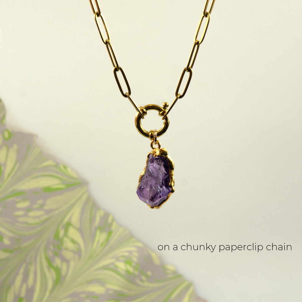 A gold paperclip chain necklace with a ring clasp holding a raw Amethyst pendant.