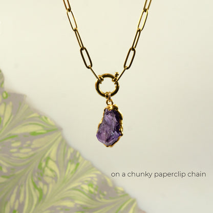 A gold paperclip chain necklace with a ring clasp holding a raw Amethyst pendant.