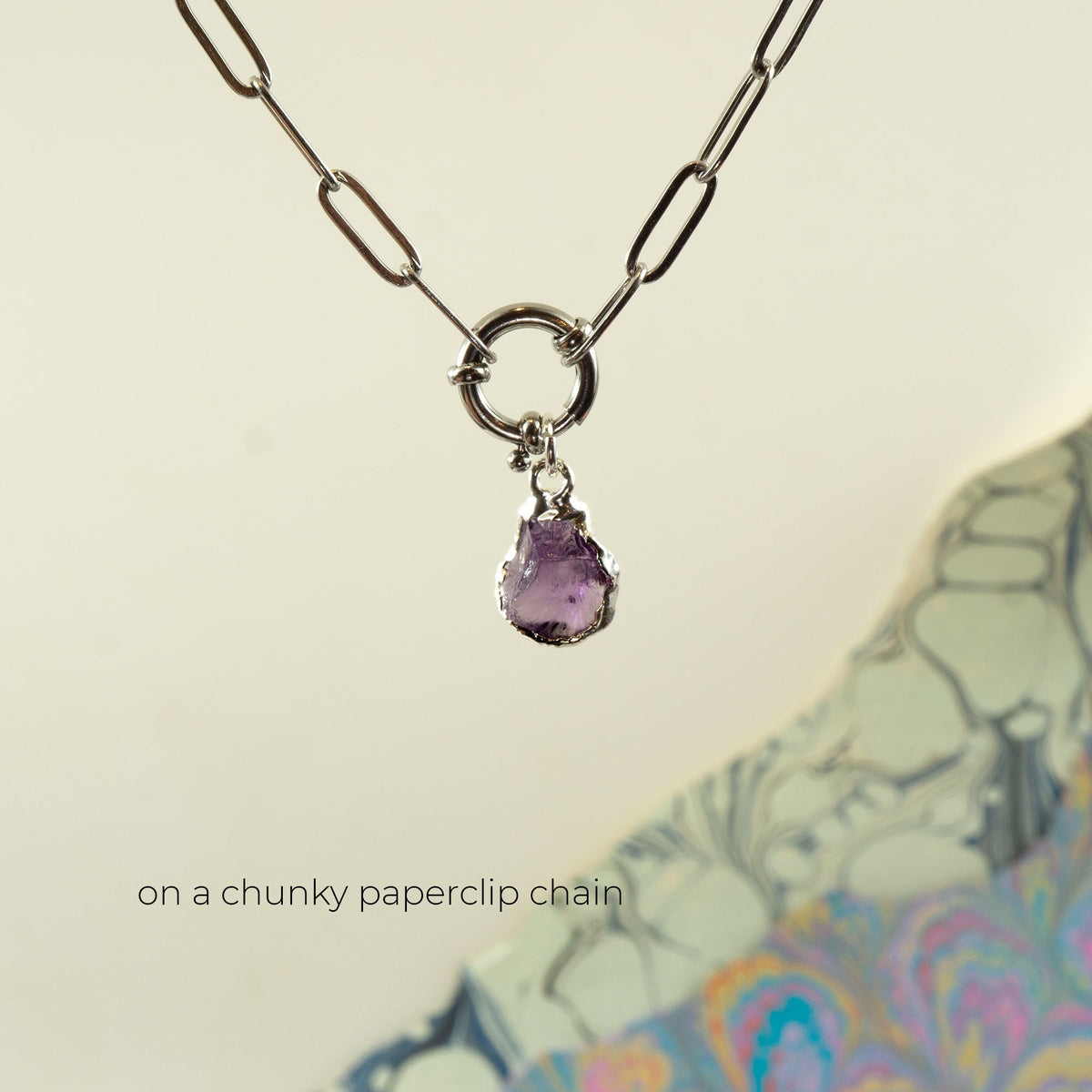 A silver paperclip chain necklace with a small ring clasp holding a teardrop-shaped purple Amethyst gemstone pendant.