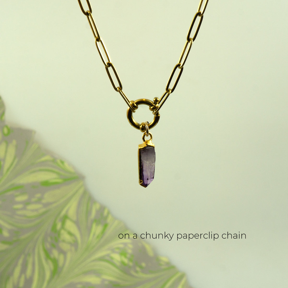 A gold chunky paperclip chain necklace with a small gold ring clasp holding a polished purple crystal pendant