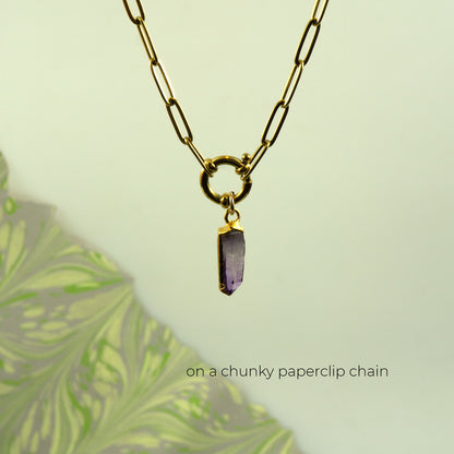 A gold chunky paperclip chain necklace with a small gold ring clasp holding a polished purple crystal pendant
