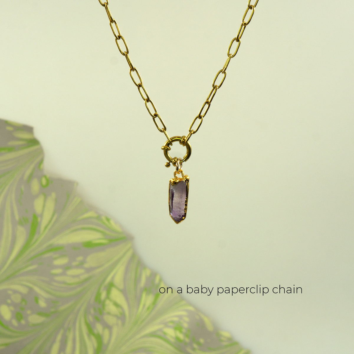 A gold paperclip chain necklace with a small gold ring clasp holding a rectangular purple amethyst crystal pendant