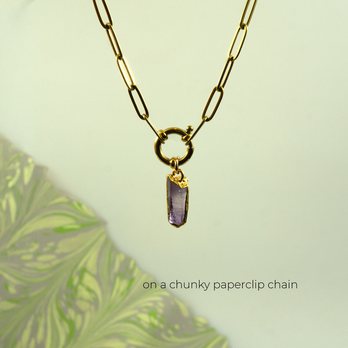 A gold paperclip chain necklace with a gold ring clasp and a small raw purple crystal pendant