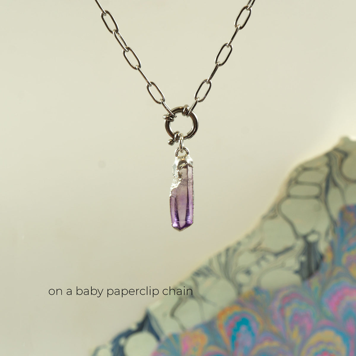 A silver paperclip chain necklace with a small silver ring and a dangling faceted purple crystal pendant