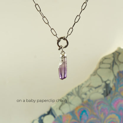 A silver paperclip chain necklace with a small silver ring and a dangling faceted purple crystal pendant