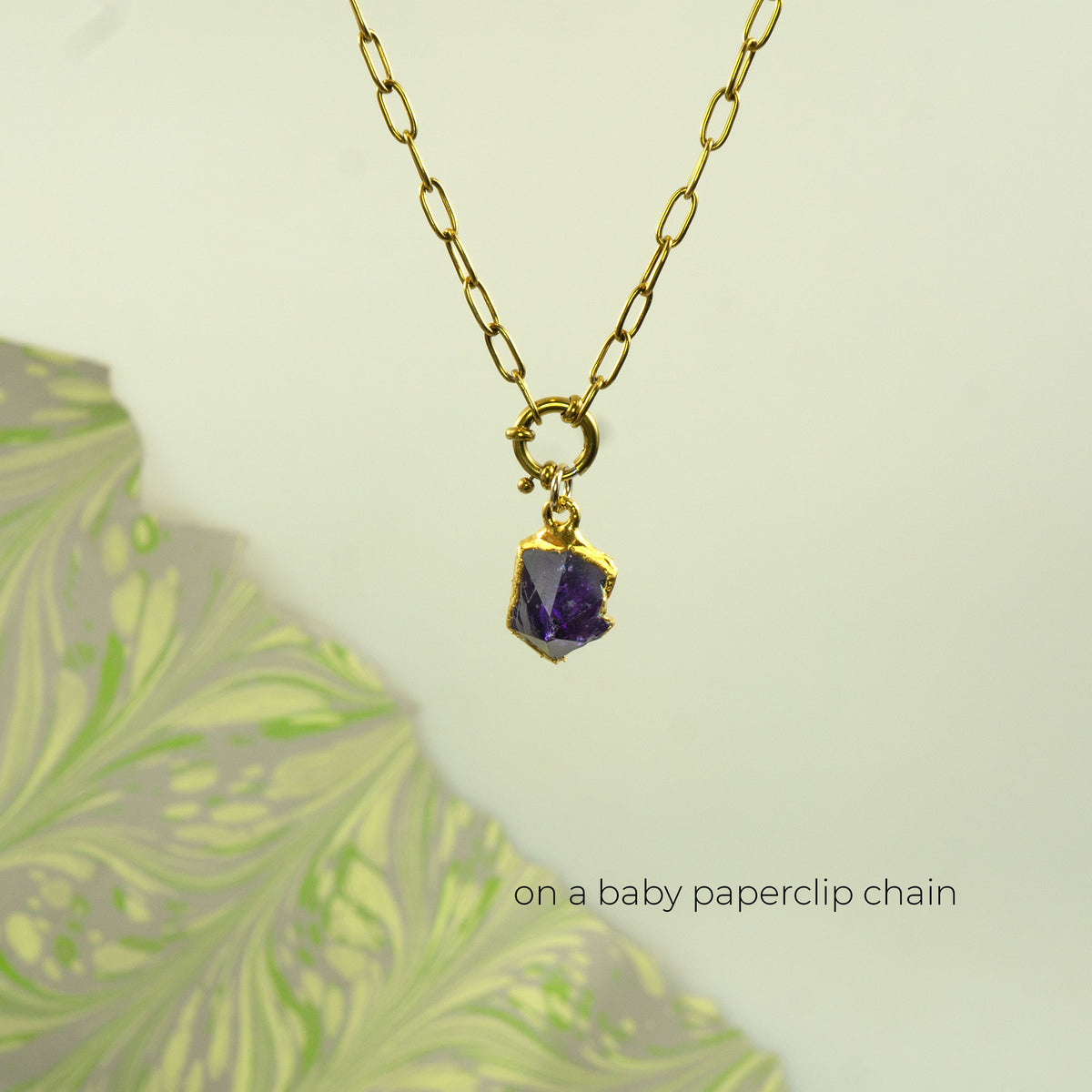 A gold paperclip chain necklace with a small gold clasp holding a faceted purple amethyst gemstone pendant,