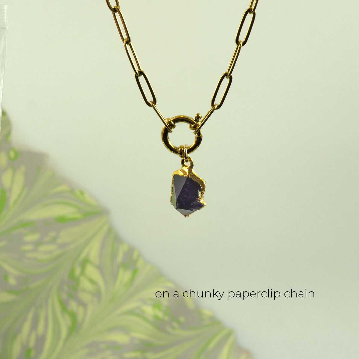A gold paperclip chain necklace with a small gold ring and a raw, rough purple gemstone pendant