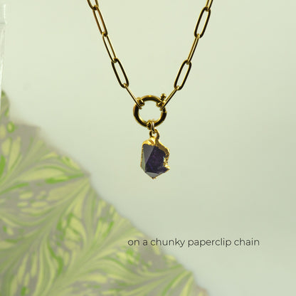 A gold paperclip chain necklace with a small gold ring and a raw, rough purple gemstone pendant