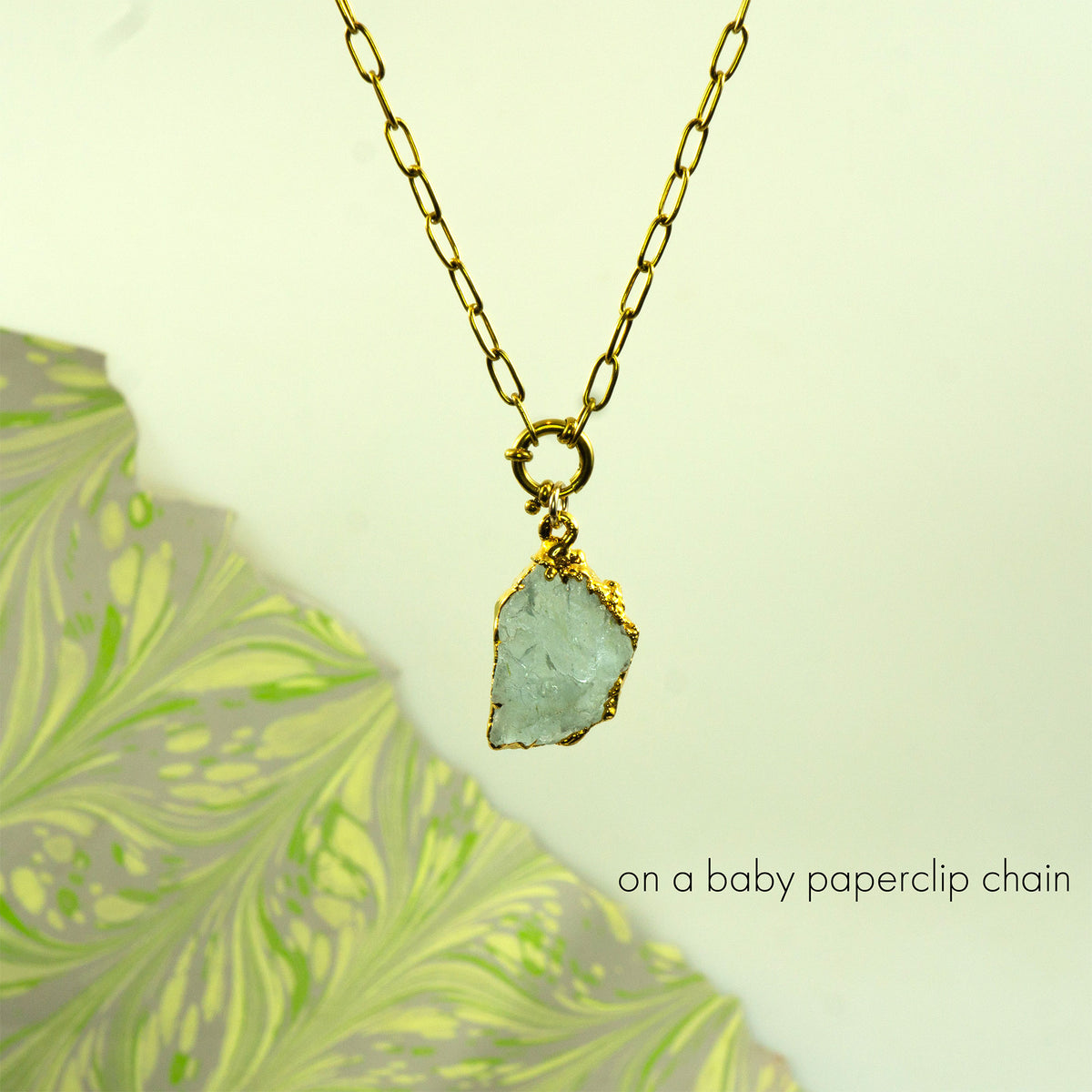 A gold paperclip chain necklace with a raw, light blue-green aquamarine gemstone pendant