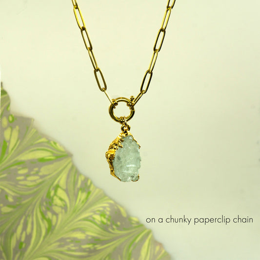 A gold necklace with a chunky paperclip chain and a raw, rough-cut light blue aquamarine gemstone pendant