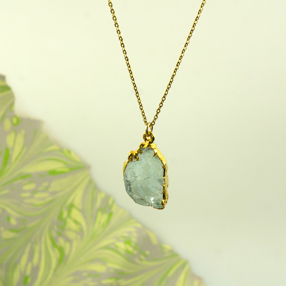 A gold chain necklace with a raw, irregular light blue-green aquamarine gemstone pendant