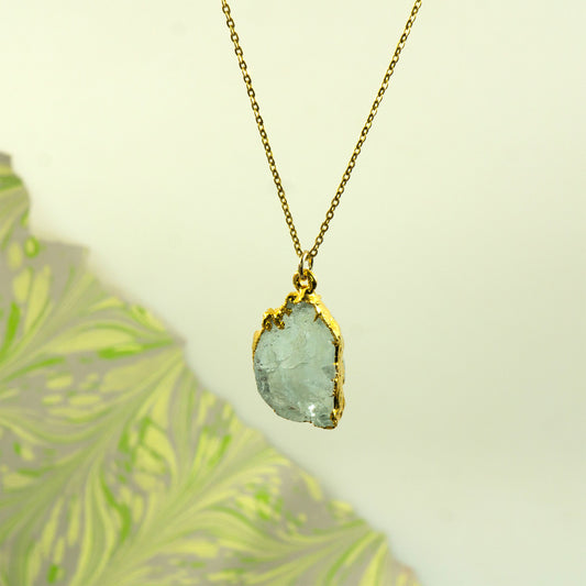 A gold chain necklace with a raw, irregular light blue-green aquamarine gemstone pendant