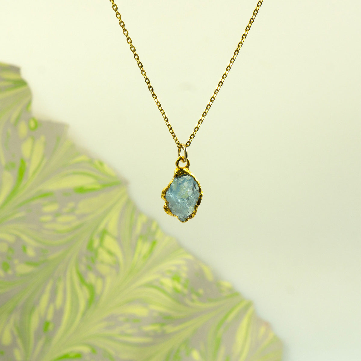 A gold chain necklace with an irregular, raw-cut aquamarine gemstone pendant in a gold bezel setting,
