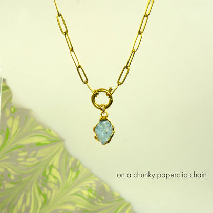 A gold necklace with a chunky paperclip chain and a small light aquamarine raw stone pendant