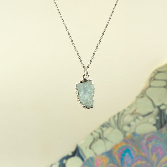 A silver chain necklace with a raw, polished light blue aquamarine gemstone pendant