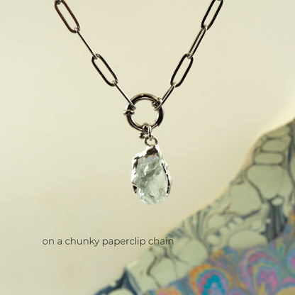 A silver paperclip chain necklace with a raw, irregular-cut aquamarine gemstone pendant